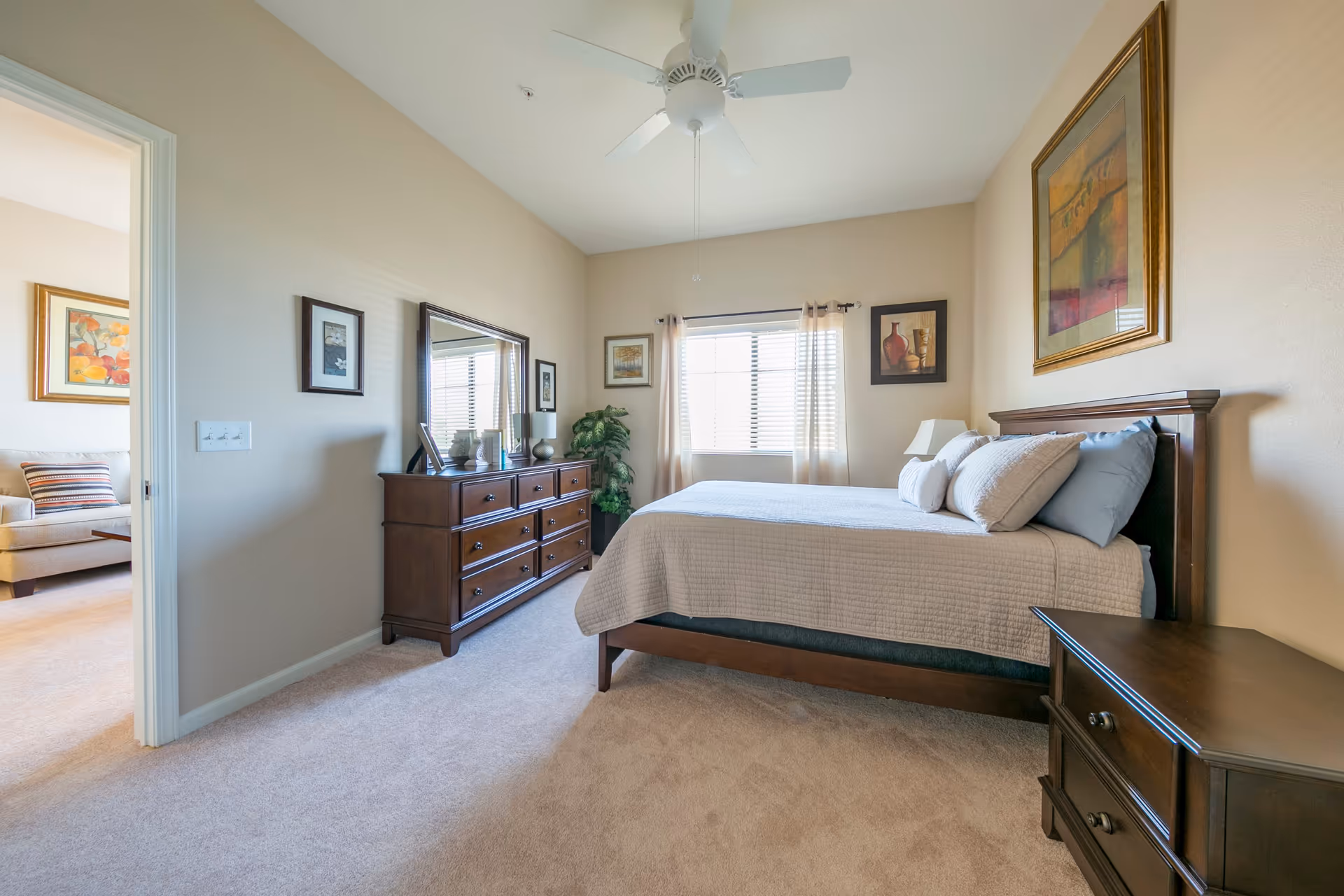 A bright bedroom with a large bed covered in a beige quilt and multiple pillows. There is a wooden dresser with a mirror and decorative items on top, a nightstand, and framed artwork on the walls. A window with light curtains lets in natural light, and a ceiling fan is mounted above. An open doorway leads to a living area with a beige armchair and a colorful pillow.