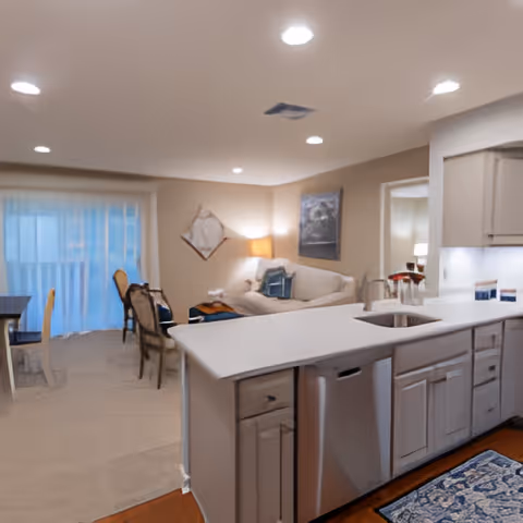 Open-concept living space featuring a kitchen with white cabinetry and a countertop island with a sink, adjacent to a living room area with a white sofa, a lamp, and wall art. In the background, there is a dining area with a table and chairs near a large window with sheer curtains.