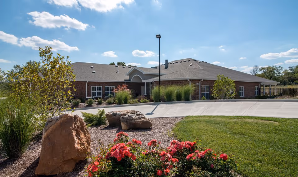 Exterior view of a single-story brick building with a gray roof under a blue sky with scattered clouds. The foreground features a landscaped area with large rocks, green shrubs, and blooming red flowers, along with a curved concrete driveway leading to the building entrance.