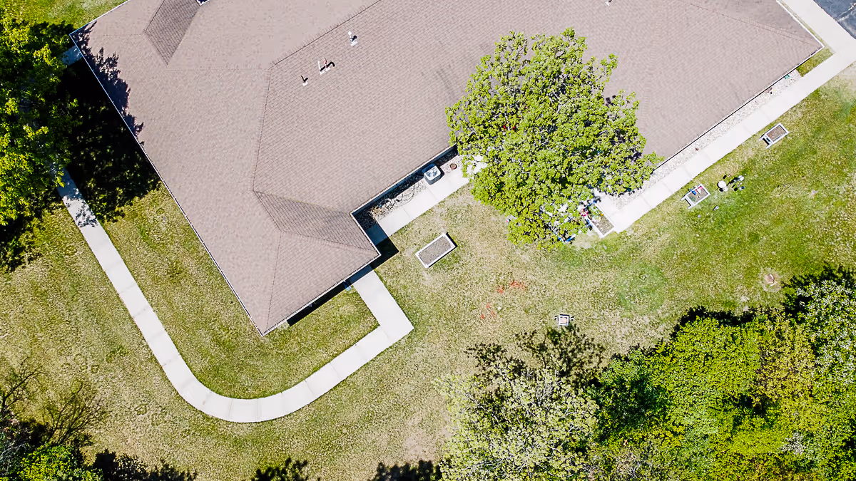 Aerial view of a senior living facility building with a brown roof, surrounded by green grass, trees, and a curved concrete walkway.