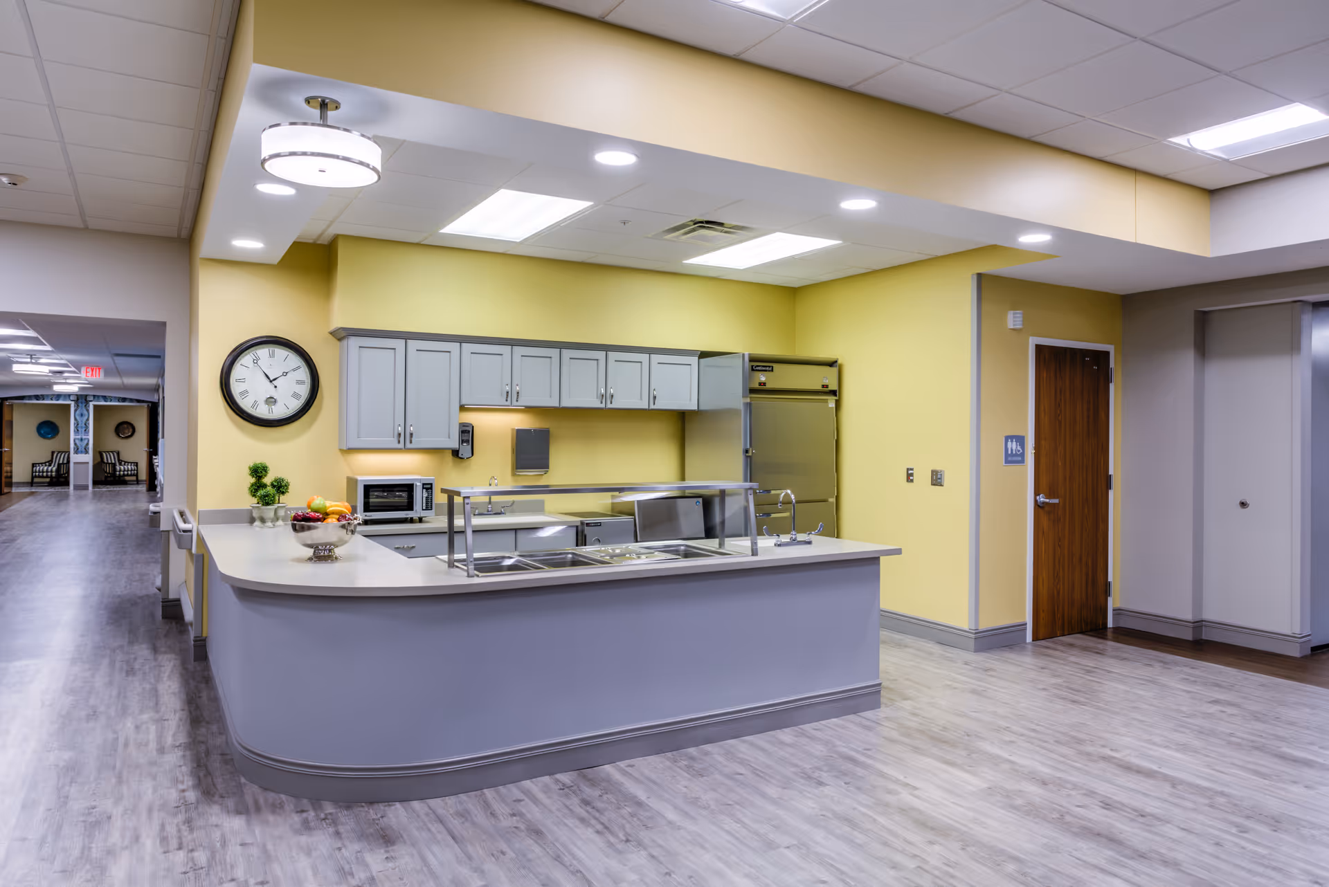 A clean and modern kitchen area in a nursing and rehabilitation community with light gray cabinets, a large stainless steel refrigerator, a microwave, a clock on the wall, and a counter with a fruit bowl and a small plant. The kitchen is open to a hallway with wood flooring and yellow walls.