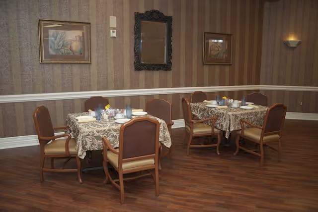 Two dining tables with patterned tablecloths set with plates, cups, and glasses in a room with wooden flooring and striped wallpaper. Each table is surrounded by four cushioned chairs. The walls are decorated with framed artwork and a decorative mirror.