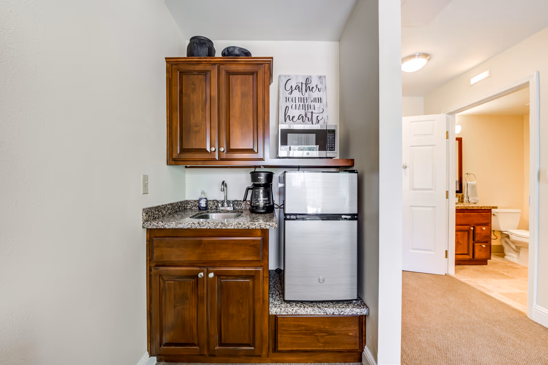 A small kitchenette area with wooden cabinets, a granite countertop, a sink, a coffee maker, a microwave, and a mini refrigerator. On the wall above the microwave is a decorative sign that reads 'Gather together with grateful hearts.' To the right, an open door reveals a bathroom with a toilet, a vanity with a granite countertop, and a mirror.