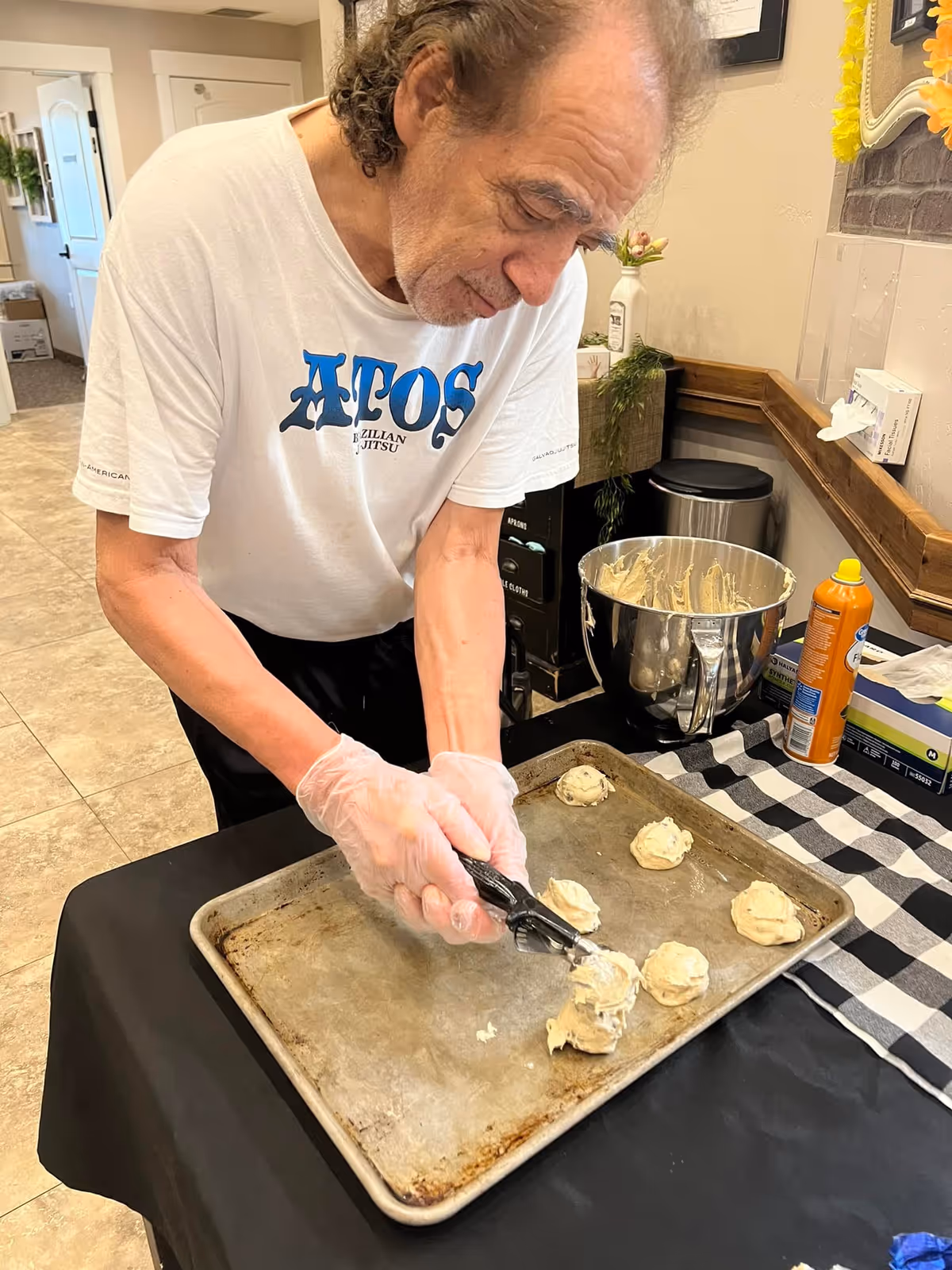 An elderly man wearing a white T-shirt and gloves is scooping cookie dough onto a baking sheet in a kitchen or food preparation area. There is a large mixing bowl with dough, a can of cooking spray, and a checkered cloth on the table. The background shows a hallway and some wall decorations.