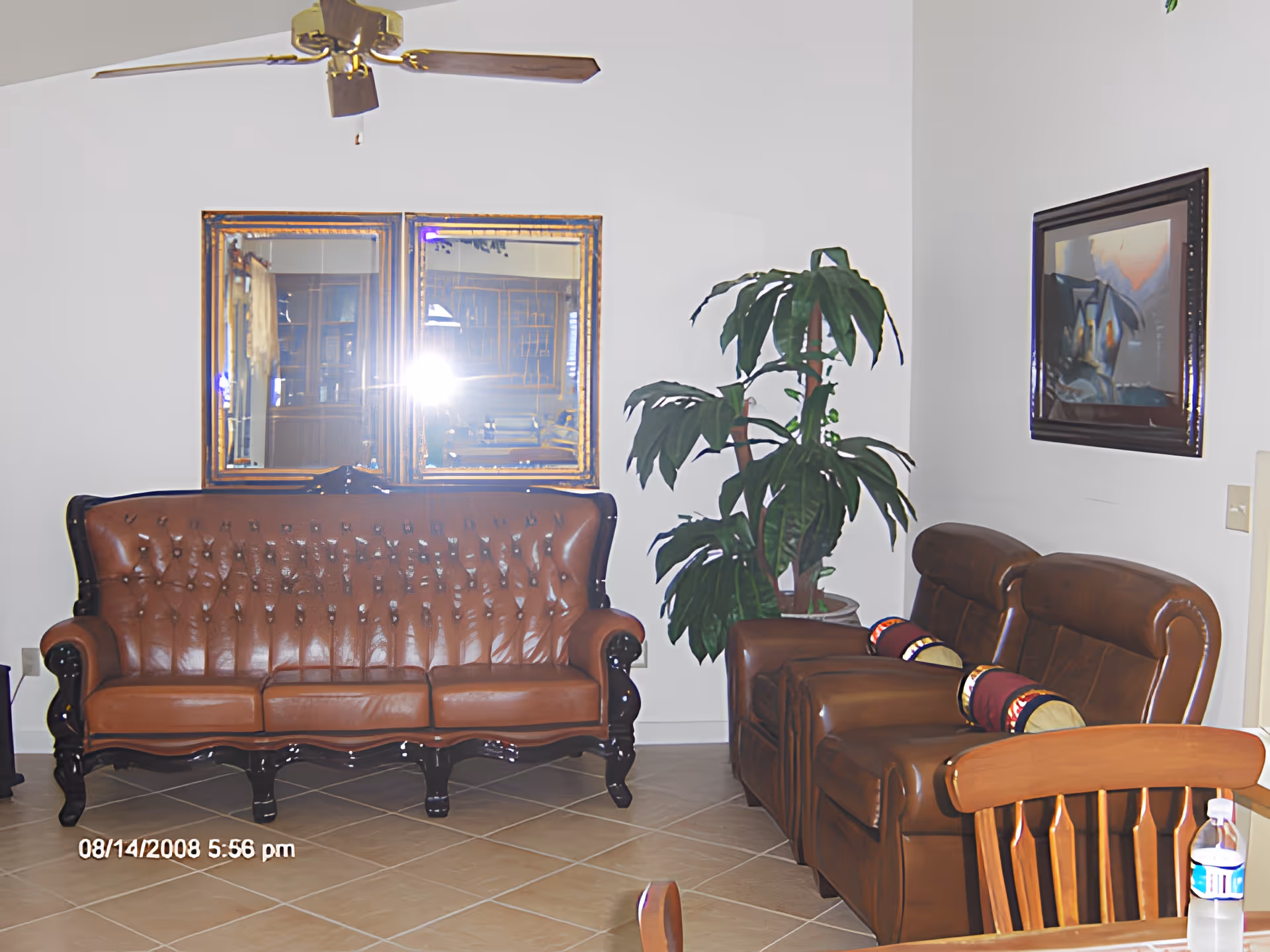 Living room with a brown tufted leather sofa and matching recliners, a potted plant, two wall mirrors, a ceiling fan, and a dining chair with a water bottle.