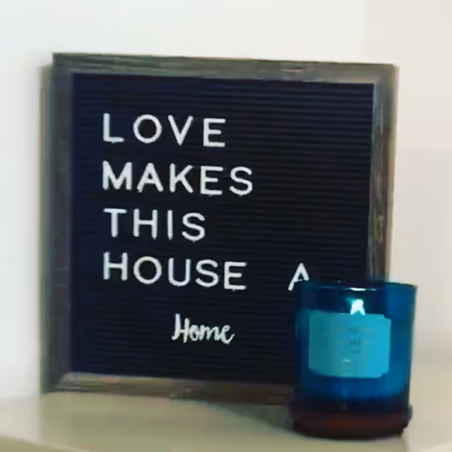 A decorative black felt letter board with a wooden frame displaying the message 'LOVE MAKES THIS HOUSE A Home' placed on a light-colored surface next to a blue glass candle holder.