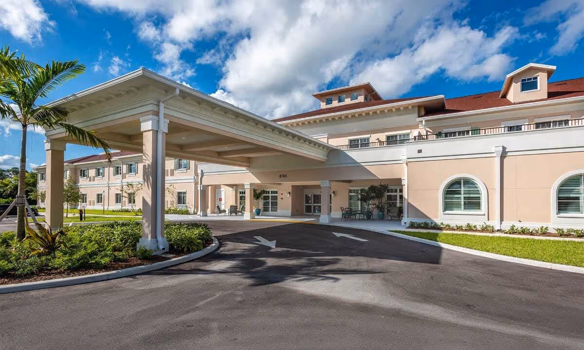 Exterior front view of a senior living facility with a covered driveway entrance, beige walls, red roof, palm trees, and a clear blue sky with some clouds.