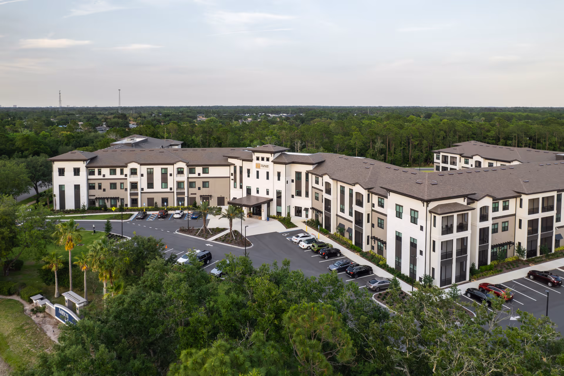Aerial view of The Gallery at Port Orange, a large three-story senior living facility surrounded by trees and greenery, with a parking lot in front and a clear sky above.