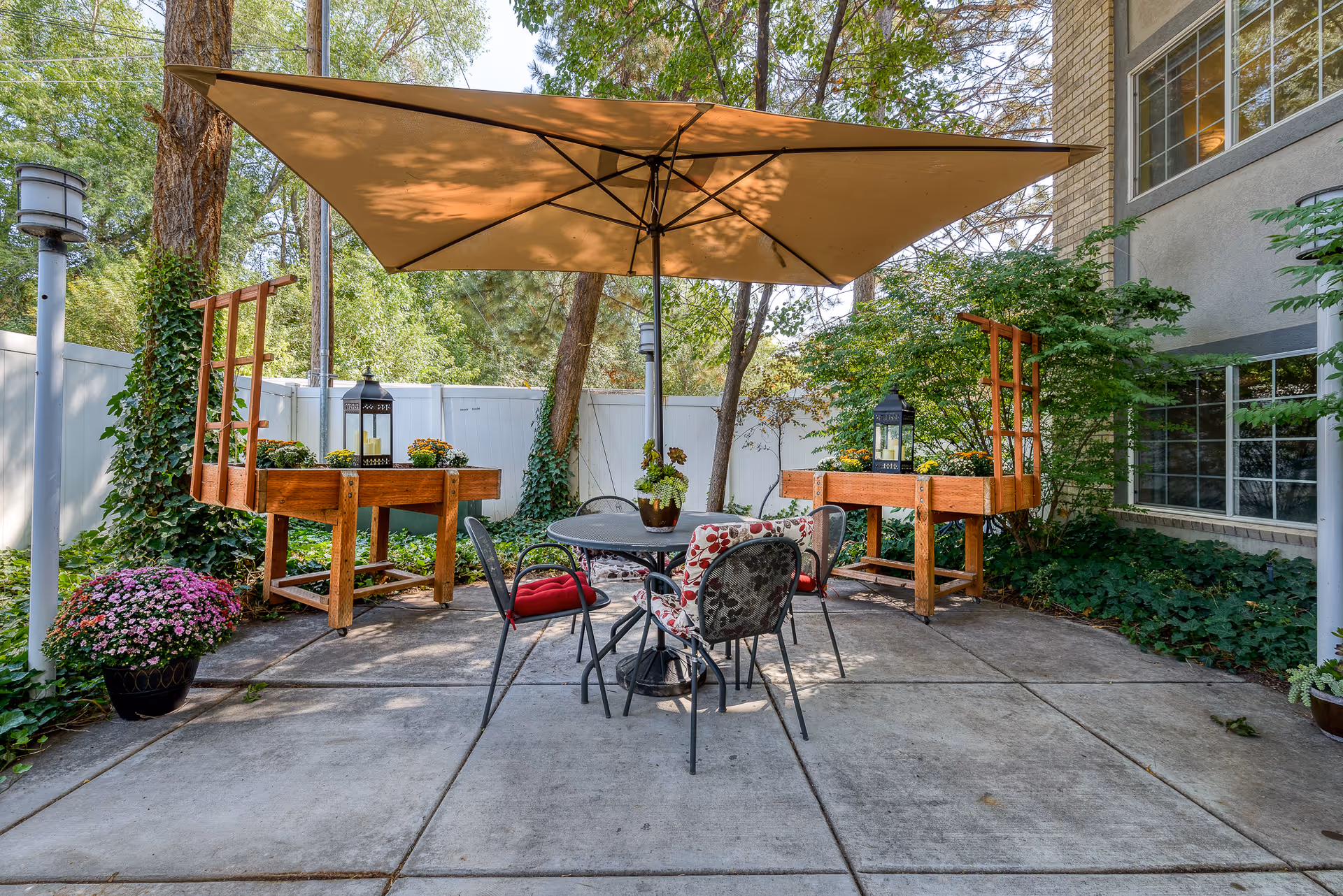 Outdoor patio area with a round metal table and four chairs, two with red cushions and one with a floral cushion. A large beige umbrella provides shade over the table. Two wooden planter stands with flowers and lanterns are positioned against a white fence, surrounded by greenery and trees.