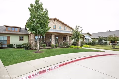 Exterior view of a single-story assisted living facility with a stone and wood facade, a covered porch with seating, green lawn, and trees in front. The driveway has a red curb marked with 'FIRE LANE NO PARKING'.