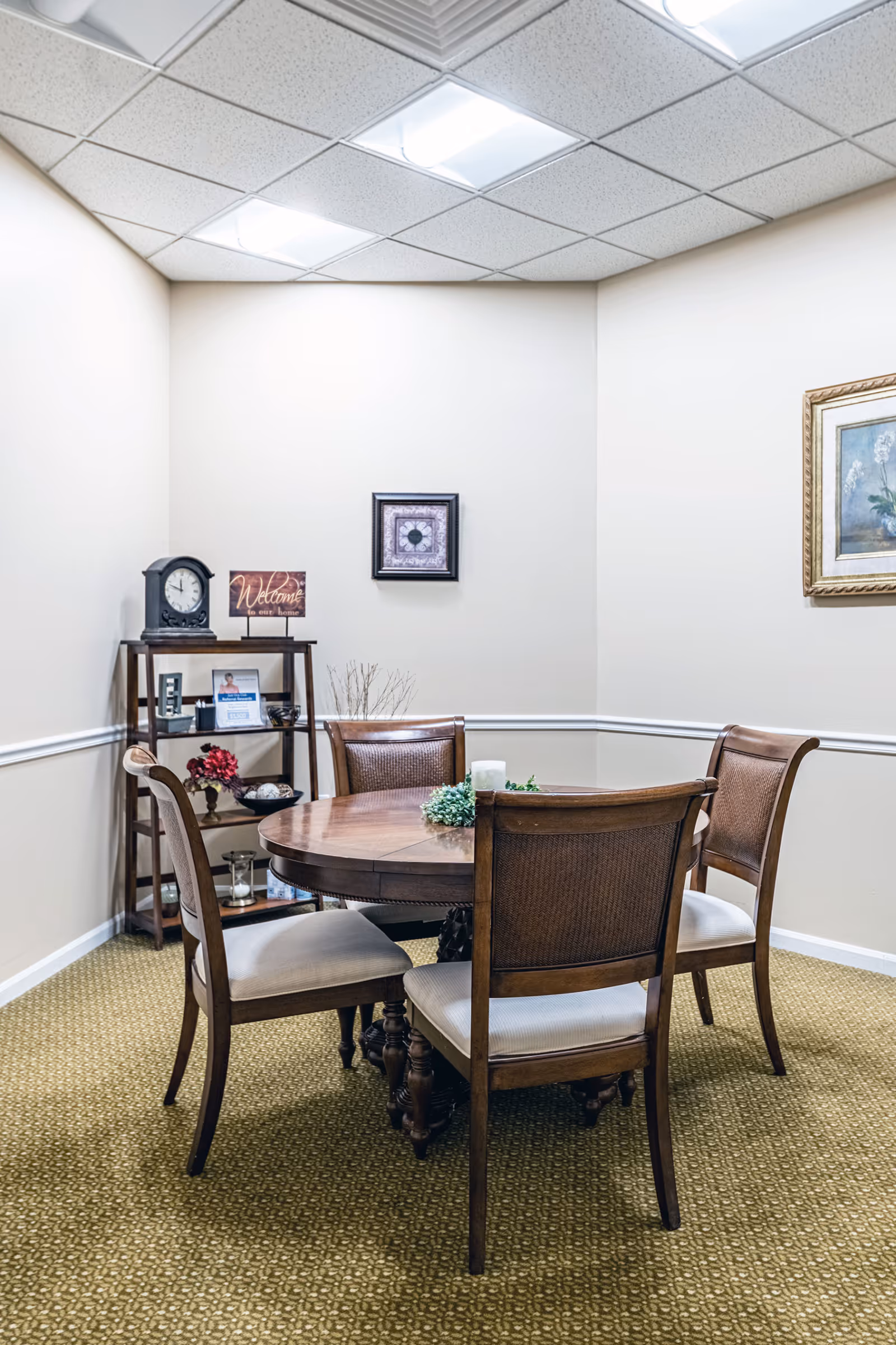 A small dining area with a round wooden table surrounded by four wooden chairs with cushioned seats. In the corner, there is a wooden shelving unit holding a clock, a welcome sign, and decorative items. The walls are light-colored with framed artwork, and the ceiling has recessed lighting panels.
