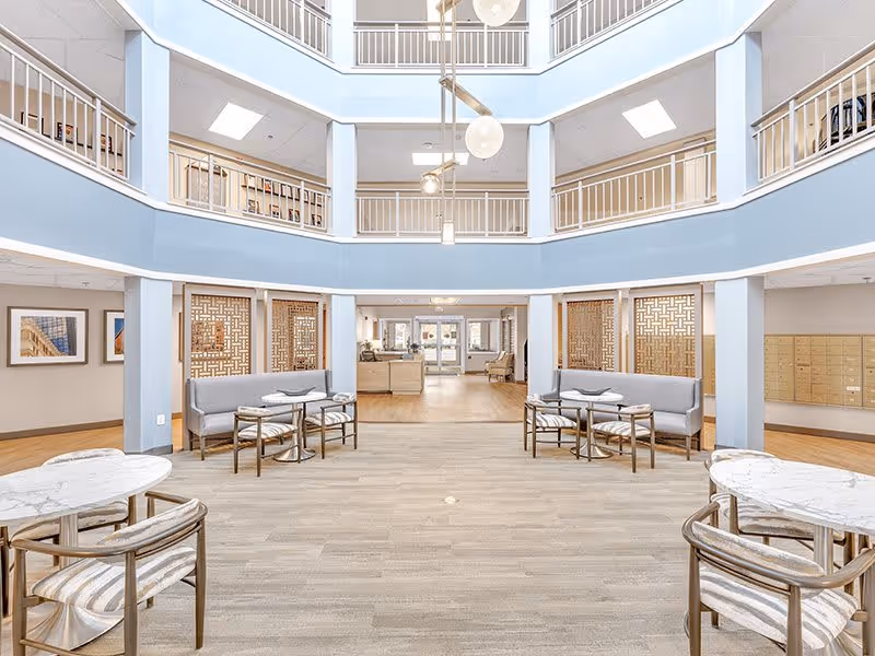 Bright and spacious interior atrium of a senior living facility with light blue walls and white railings on the upper floors. The ground floor features several seating areas with gray cushioned benches and round marble-top tables with chairs. The space has wooden flooring and decorative wooden panels on the walls, with mailboxes visible on the right side and framed artwork on the left.