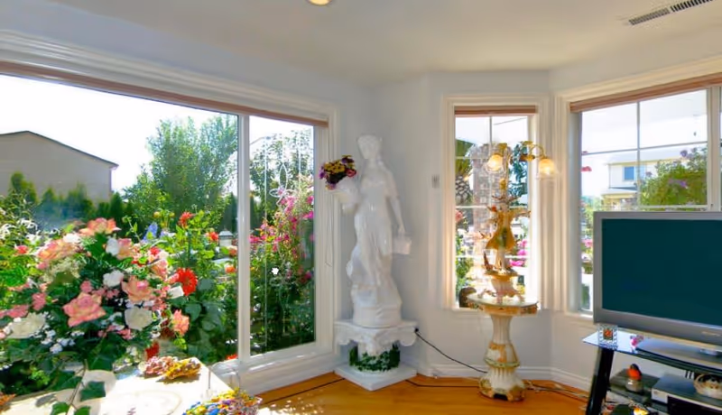 A bright living room with large windows showcasing vibrant flowers outside. A white statue stands elegantly beside a decorative table, and a television is positioned in the corner.
