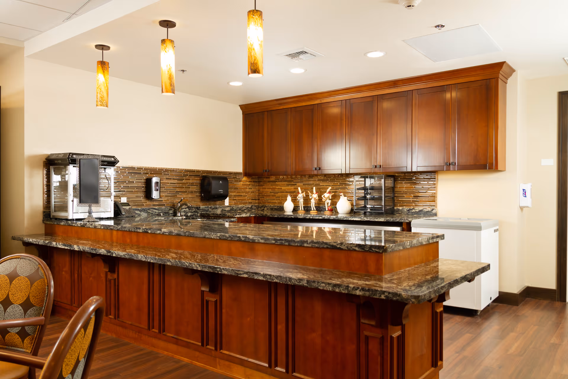 Interior view of a kitchen area with a large granite countertop island, wooden cabinets, three pendant lights hanging from the ceiling, a popcorn machine on the counter, and decorative items on the back counter. There are also chairs with patterned upholstery partially visible in the foreground.