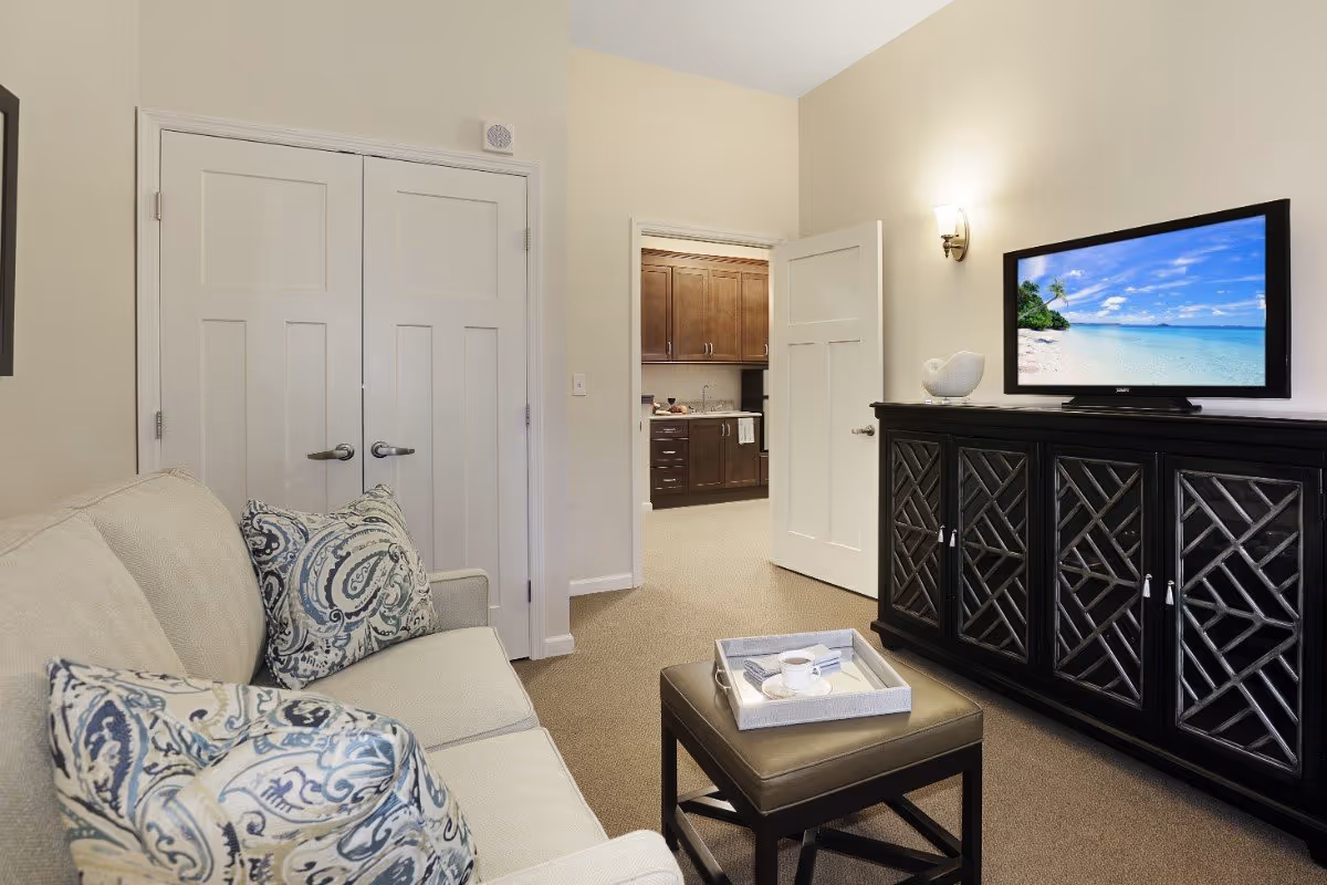 Cozy living room with a beige sofa and patterned pillows, a leather ottoman with a tray, and a TV on a dark cabinet facing an open doorway to a kitchen.