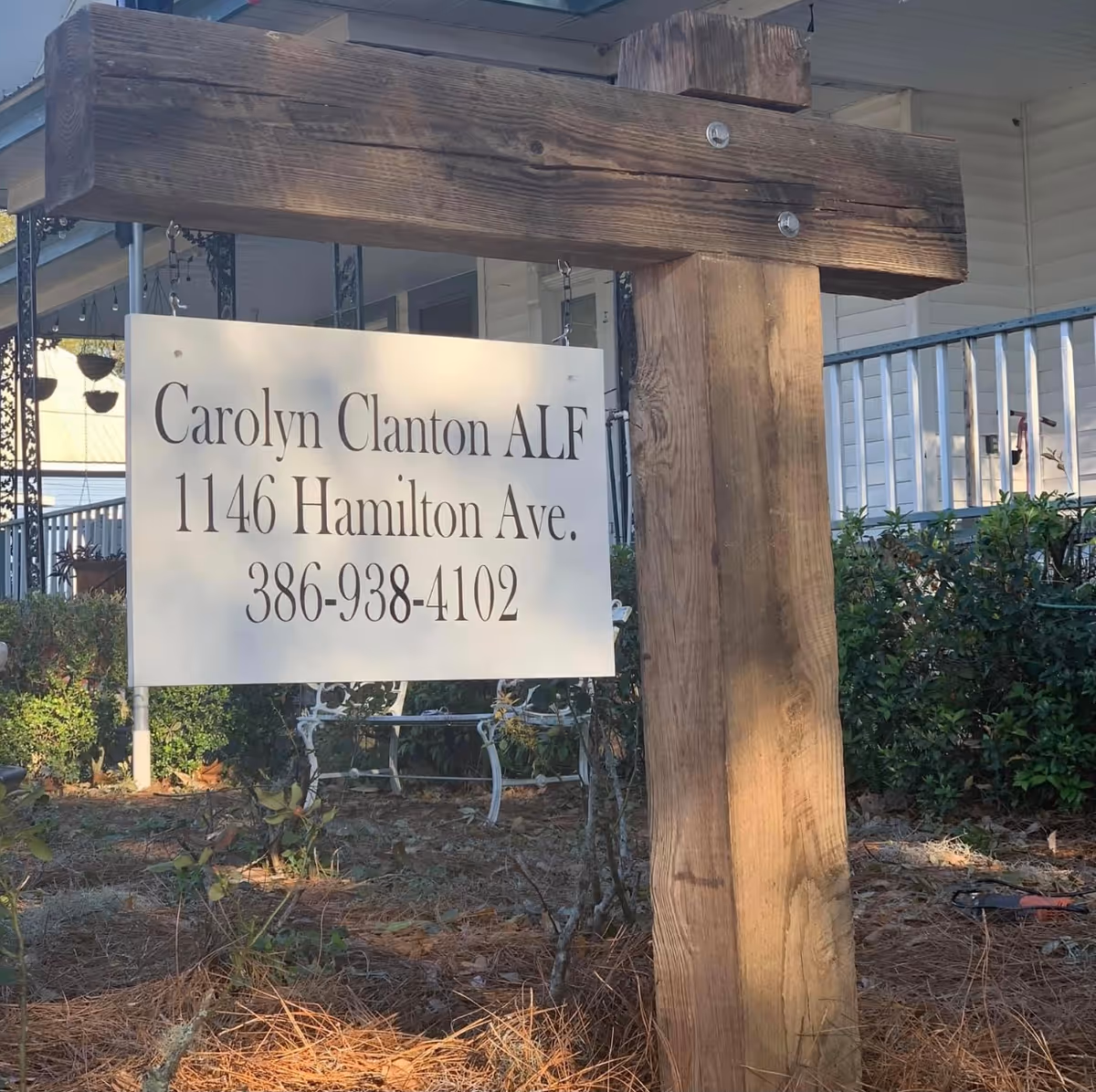 A wooden post with a hanging white sign displaying the text 'Carolyn Clanton ALF 1146 Hamilton Ave. 386-938-4102' in front of a house with a porch and some bushes.