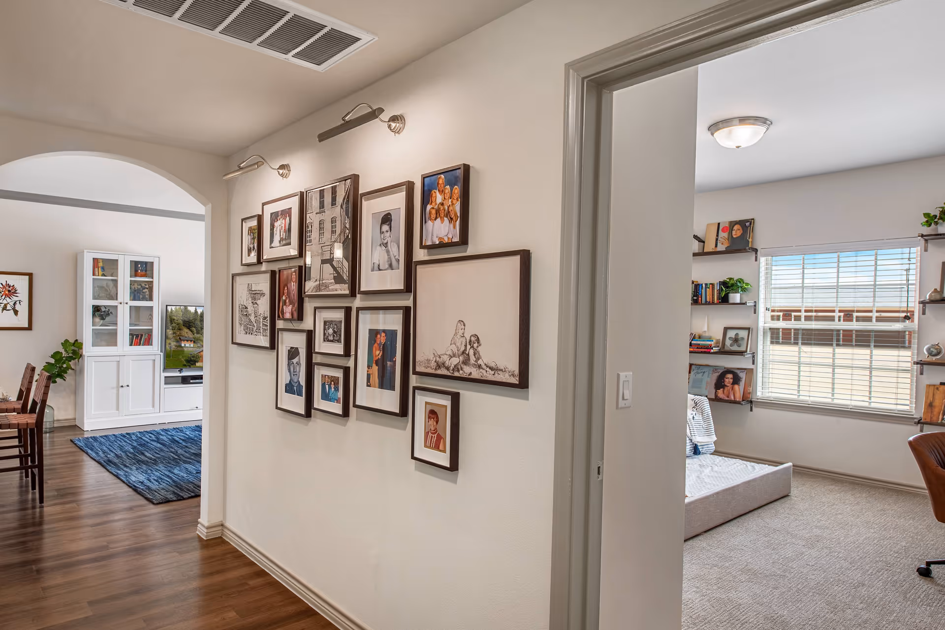 Interior view of a senior living facility hallway with a gallery wall of framed photographs and artwork. To the left, there is an arched doorway leading to a dining area with a wooden floor, chairs, a blue rug, and a white cabinet with a TV. To the right, there is a doorway leading to a carpeted room with a window, shelves with books and plants, and a chair.