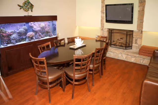 A cozy interior room with a dark wooden oval table surrounded by eight matching chairs. To the left, there is a large built-in aquarium filled with coral and fish. On the right side, there is a stone fireplace with a mounted flat-screen TV above it. The floor is wooden, and there are cushioned bench seats along the walls.