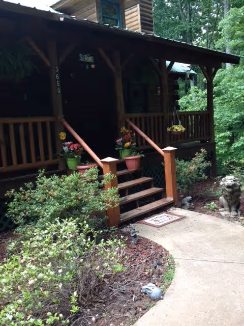 Front porch of a wooden cabin-style building with steps leading up to the entrance, surrounded by green shrubs and plants. There are potted flowers on the steps and a stone lion statue near the walkway.