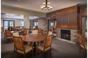 A cozy dining area in a senior living community featuring multiple round wooden tables surrounded by cushioned chairs with patterned upholstery. The room has a stone fireplace with a wooden mantle, a chandelier hanging from the ceiling, and large windows allowing natural light to enter.