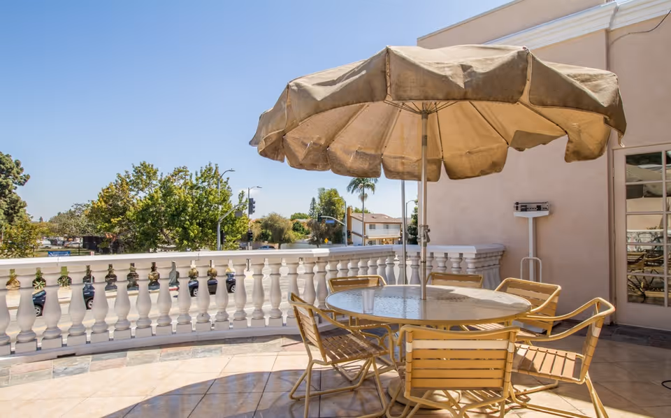 Outdoor patio area with a round glass table surrounded by six wooden chairs. A large beige umbrella is positioned in the center of the table providing shade. The patio has a white balustrade railing and overlooks a street with trees and buildings in the background under a clear blue sky.