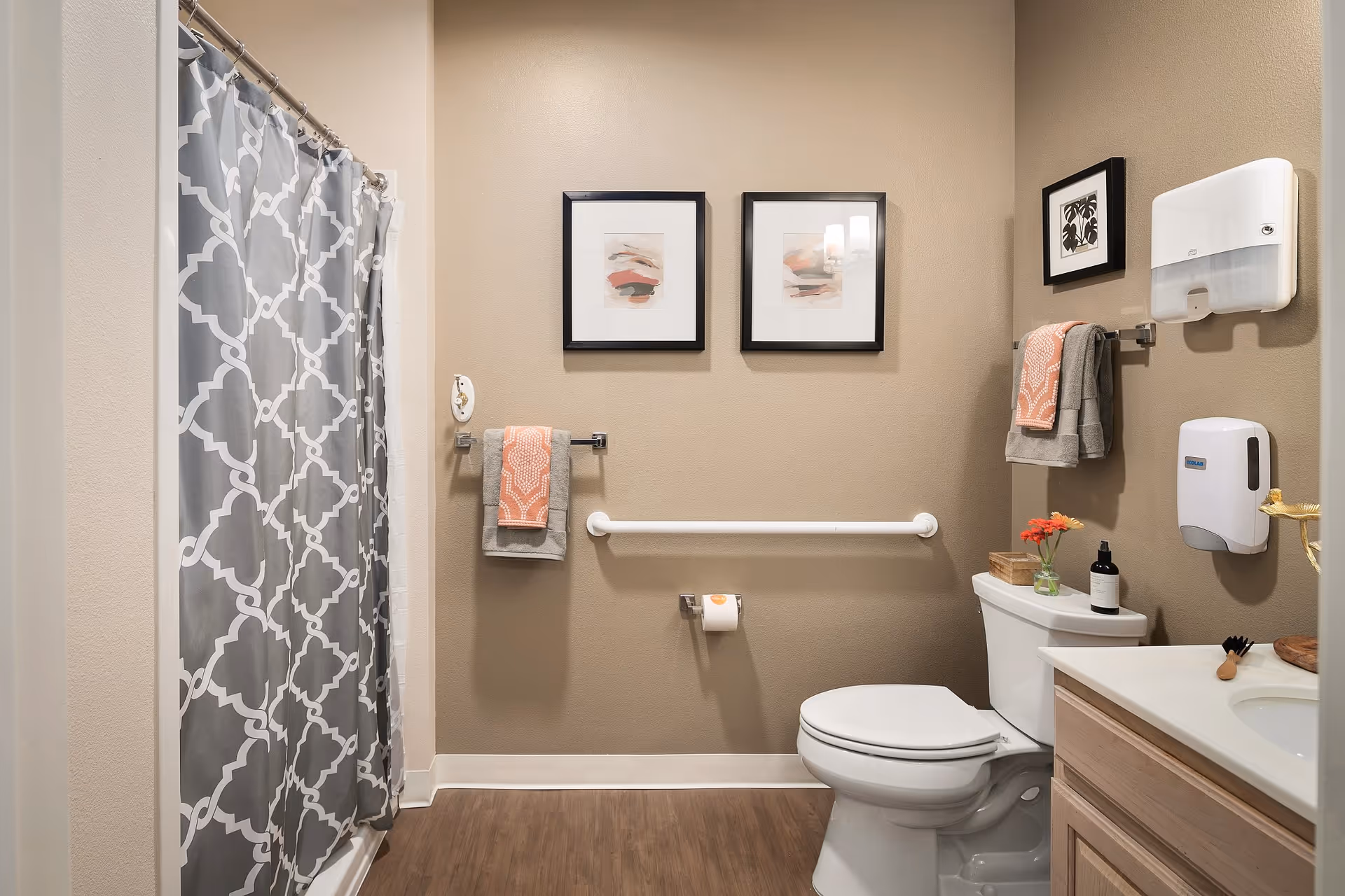 A clean and well-lit bathroom with beige walls and wood-style flooring. There is a white toilet with a small vase of flowers and a soap dispenser on the tank. A white grab bar is mounted on the wall behind the toilet, with a toilet paper holder below it. Two framed abstract art pieces hang above the grab bar. To the left, there is a shower with a gray and white patterned curtain. On the right wall, there are two towel racks holding gray and coral towels, a wall-mounted soap dispenser, and a paper towel dispenser. A wooden vanity with a white countertop and sink is partially visible on the right side.