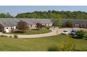 A single-story brick nursing and rehab center building with a curved driveway and a few parked cars, surrounded by green grass and trees in the background under a clear sky.