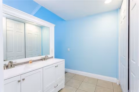 A clean bathroom interior with light blue walls, a large mirror above a white double-sink vanity with silver faucets, and tiled floor. There are white paneled doors on the right side of the room.