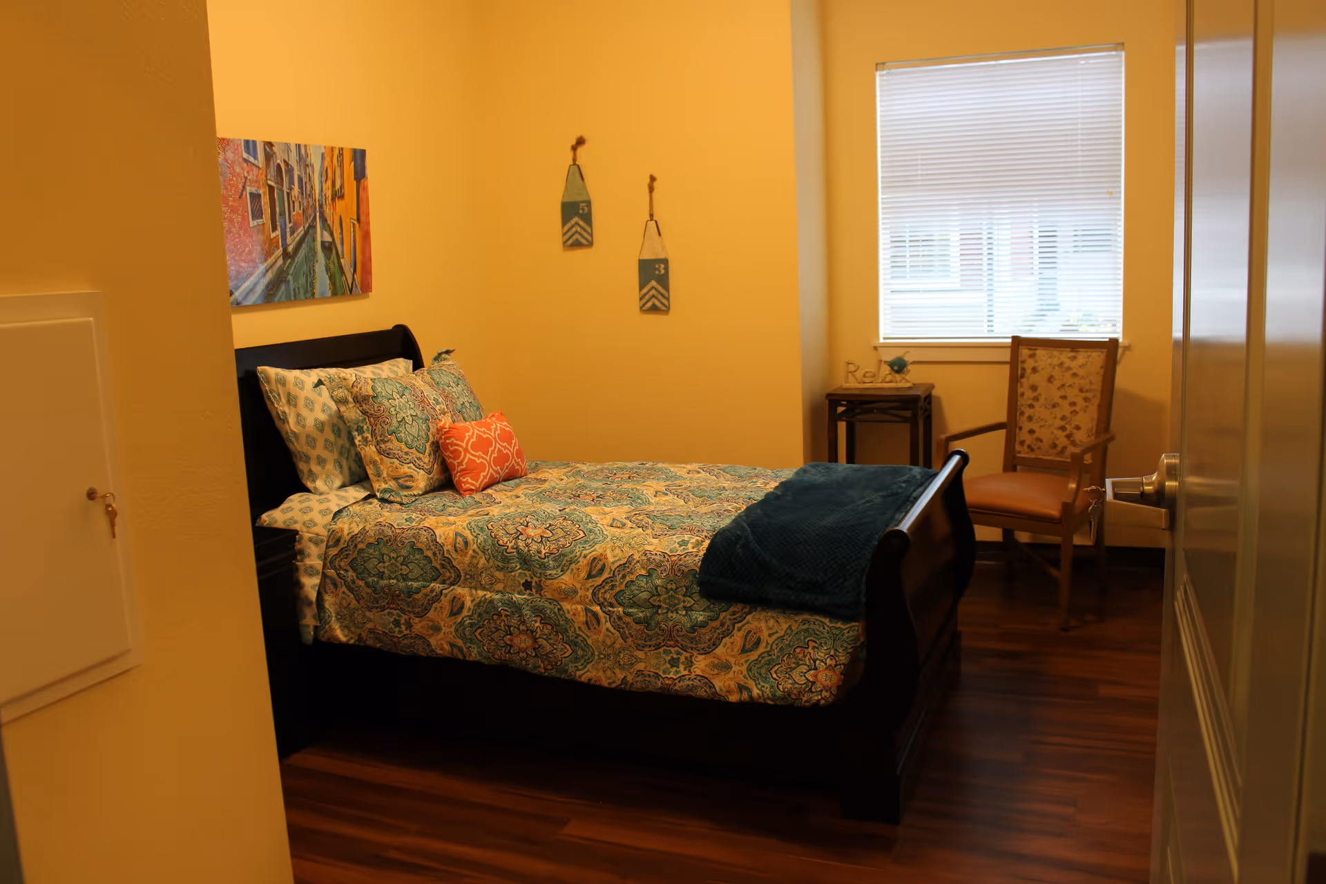 A cozy bedroom with a dark wooden bed frame and a bed made with patterned blue and green bedding, accented by a coral decorative pillow. There is a wooden chair with floral upholstery near a window with closed blinds. A small side table next to the chair holds a decorative item and a sign that says 'Relax'. The walls are painted a warm beige color and there is a colorful painting of a canal scene hanging above the bed. Two decorative hanging plaques with numbers 5 and 3 are on the wall beside the bed. The floor is a polished wood finish.