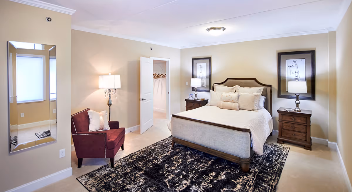 A well-lit bedroom with a large bed featuring multiple pillows and a wooden frame. There are two matching wooden nightstands on either side of the bed, each with a lamp and framed artwork above. A dark patterned rug covers the floor beneath the bed. To the left, there is a maroon armchair with a decorative pillow and a tall floor lamp beside it. A large rectangular mirror is mounted on the wall near a window with closed blinds. An open door reveals a walk-in closet with hangers inside.