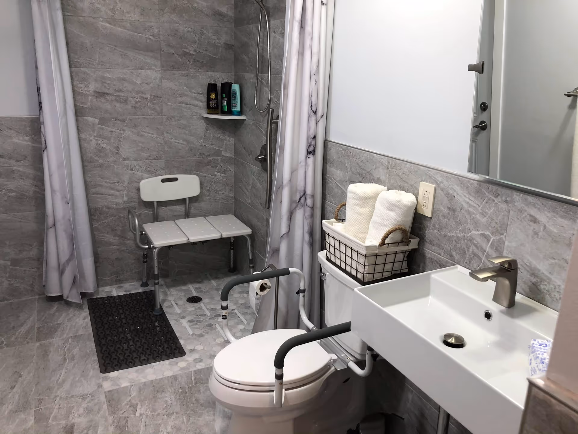 A modern accessible bathroom with gray tiled walls and floor. It features a walk-in shower with a white shower chair, a handheld showerhead, and a black non-slip mat. There is a white toilet with support rails on both sides and a basket with rolled white towels on the toilet tank. A white rectangular sink with a silver faucet is mounted on the wall next to the toilet, and a large mirror is above the sink.