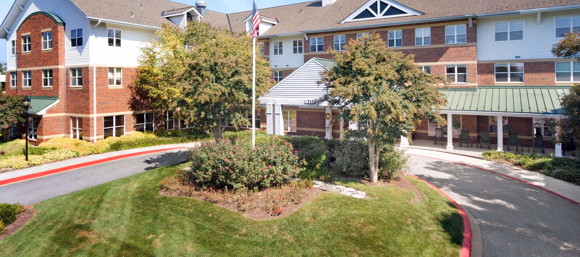 Exterior view of a senior living facility with a circular driveway, landscaped garden with bushes and trees, and an American flag on a flagpole in front of the building. The building has a brick and white siding facade with multiple windows and a green metal roof over the entrance.