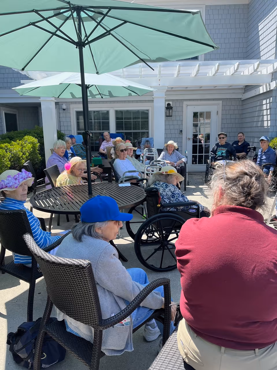 A group of elderly people sitting outdoors under large green umbrellas on a patio at a senior living facility. Some individuals are seated in wheelchairs, while others sit in chairs around tables. The background shows a building with gray siding and a white pergola.