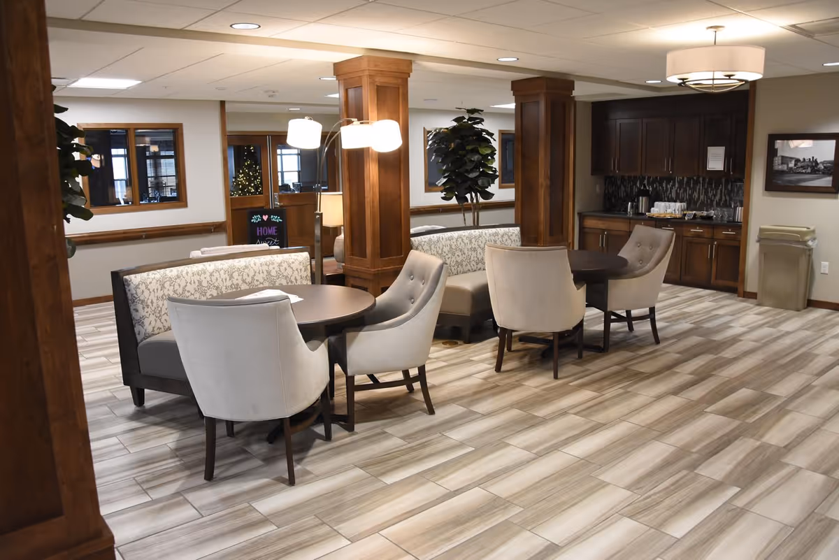 A cozy seating area in a senior living facility with round tables, cushioned chairs, and booth-style benches. The space features wooden columns, a tiled floor, warm lighting, and a small kitchenette area with cabinets and a countertop in the background.