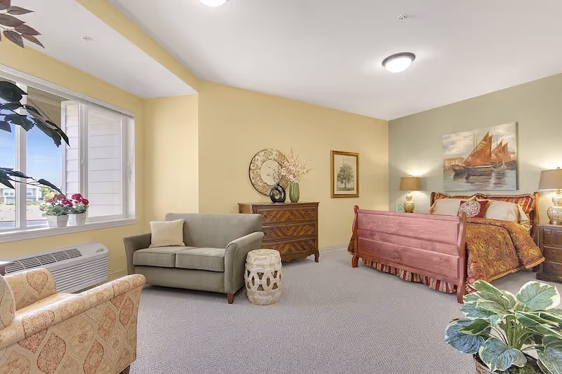 Bright studio-style suite with a wooden bed, upholstered seating, dresser, and a large window.