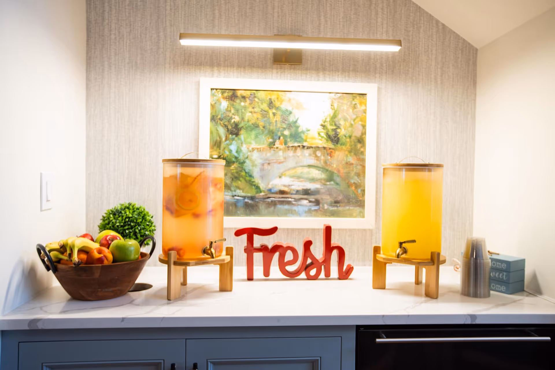 A countertop with two beverage dispensers filled with fruit-infused drinks, a wooden bowl of assorted fresh fruits, a decorative red sign that says 'Fresh', a small green plant, and a stack of plastic cups. Above the countertop is a framed painting of a stone bridge over a river surrounded by greenery, illuminated by a horizontal light fixture.