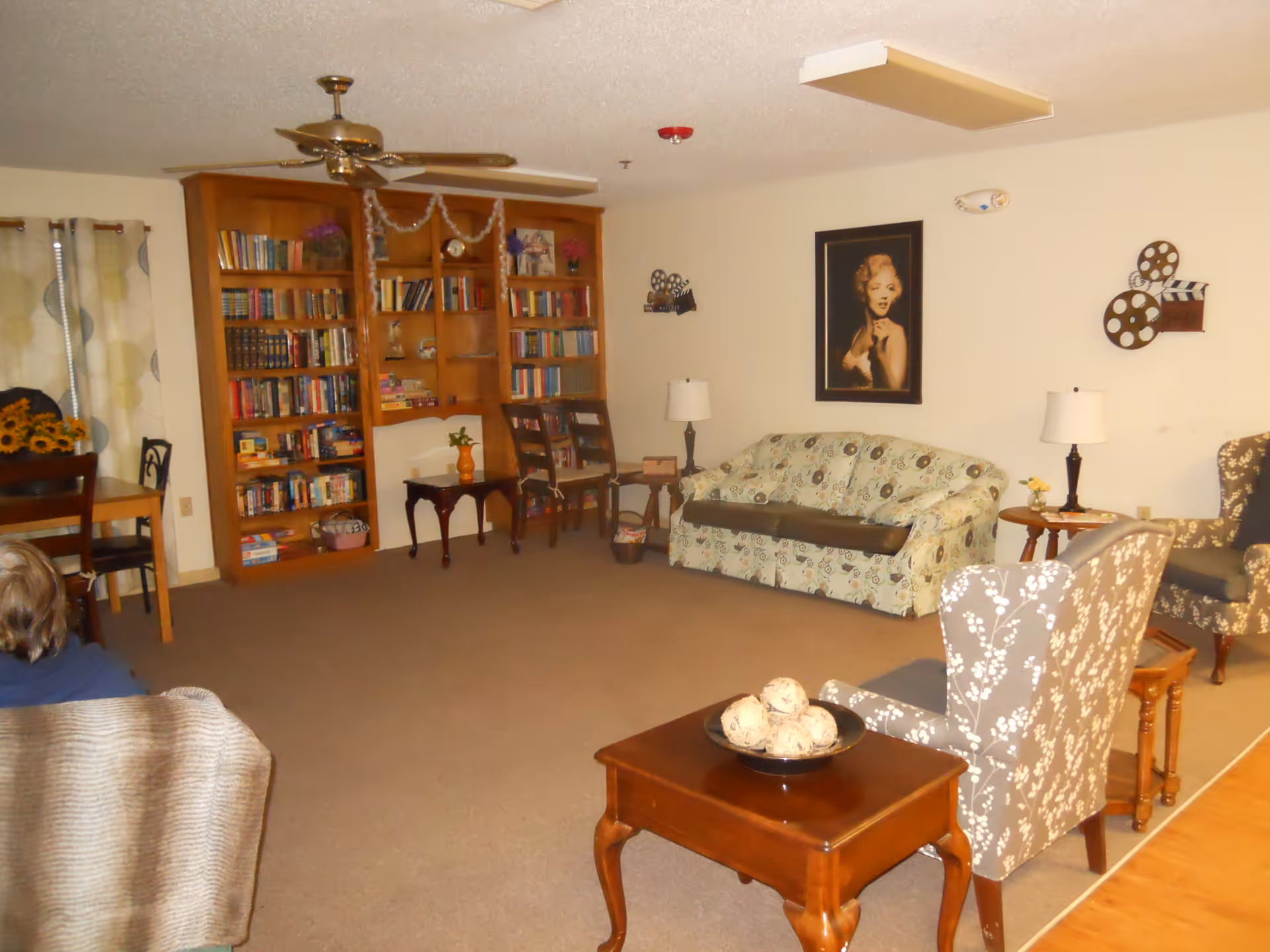 A cozy living room area with a patterned sofa, two floral armchairs, wooden side tables with lamps, a wooden coffee table with decorative balls, a large wooden bookshelf filled with books, and a framed portrait of a woman on the wall. There is also a dining table with chairs and curtains with a circular pattern on the window.