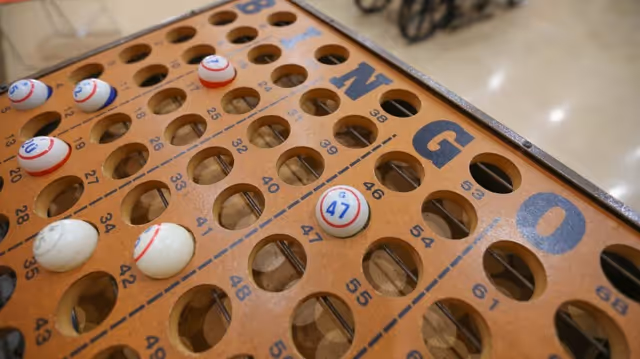 Close-up view of a wooden bingo board with numbered slots and several bingo balls placed in the holes, showing numbers like 47 and 10. The background is blurred, suggesting an indoor setting.