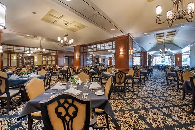 Spacious dining room with multiple tables covered in dark tablecloths, each set with white napkins, plates, glasses, and floral centerpieces. The room features ornate patterned carpeting, wooden paneling, chandeliers, and large windows allowing natural light to enter.