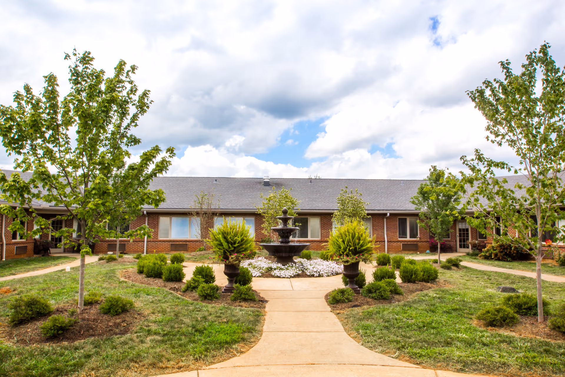 Outdoor courtyard area of a senior living facility with a central three-tiered fountain surrounded by flower beds and greenery. The courtyard is landscaped with small trees, shrubs, and a paved walkway leading to the building entrance. The building is a single-story brick structure with multiple windows under a cloudy sky.