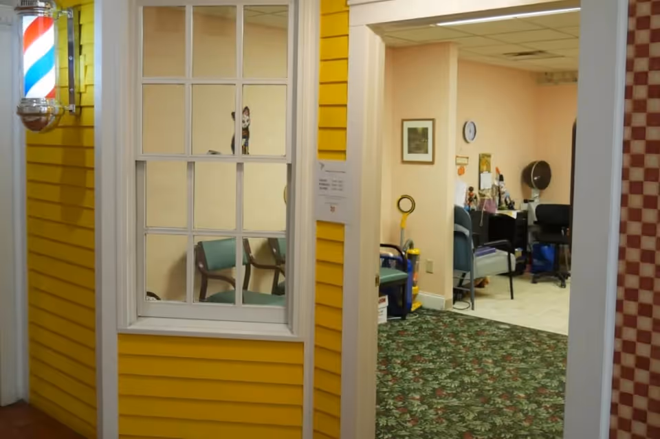 Interior view of a senior living facility showing a yellow wall with a window and a traditional barber pole light fixture. Through an open doorway, a room with chairs, a clock on the wall, framed artwork, and various office items is visible. The floor has a floral patterned carpet.