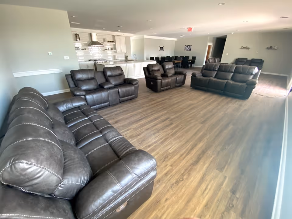 Open common living area with several dark leather recliner sofas on wood flooring and a kitchen/dining area in the background.