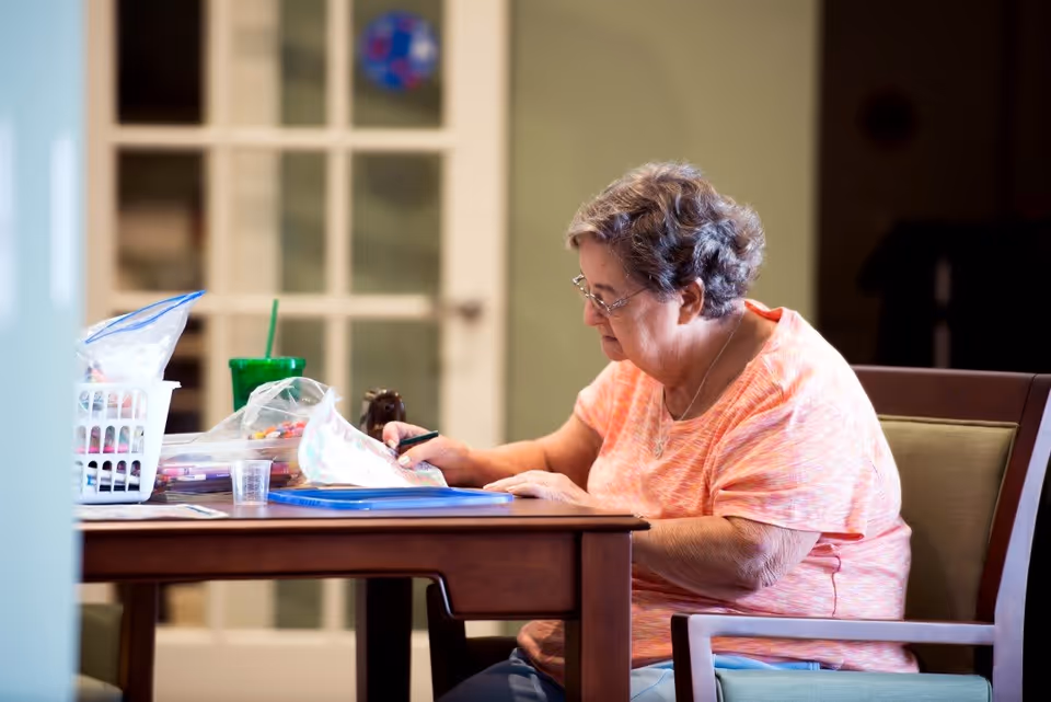 An elderly woman with short gray hair and glasses is sitting at a wooden table in a well-lit room, engaged in coloring or drawing on a piece of paper. The table has various art supplies, including markers and a green cup with a straw. Behind her is a glass-paneled door and a green wall.