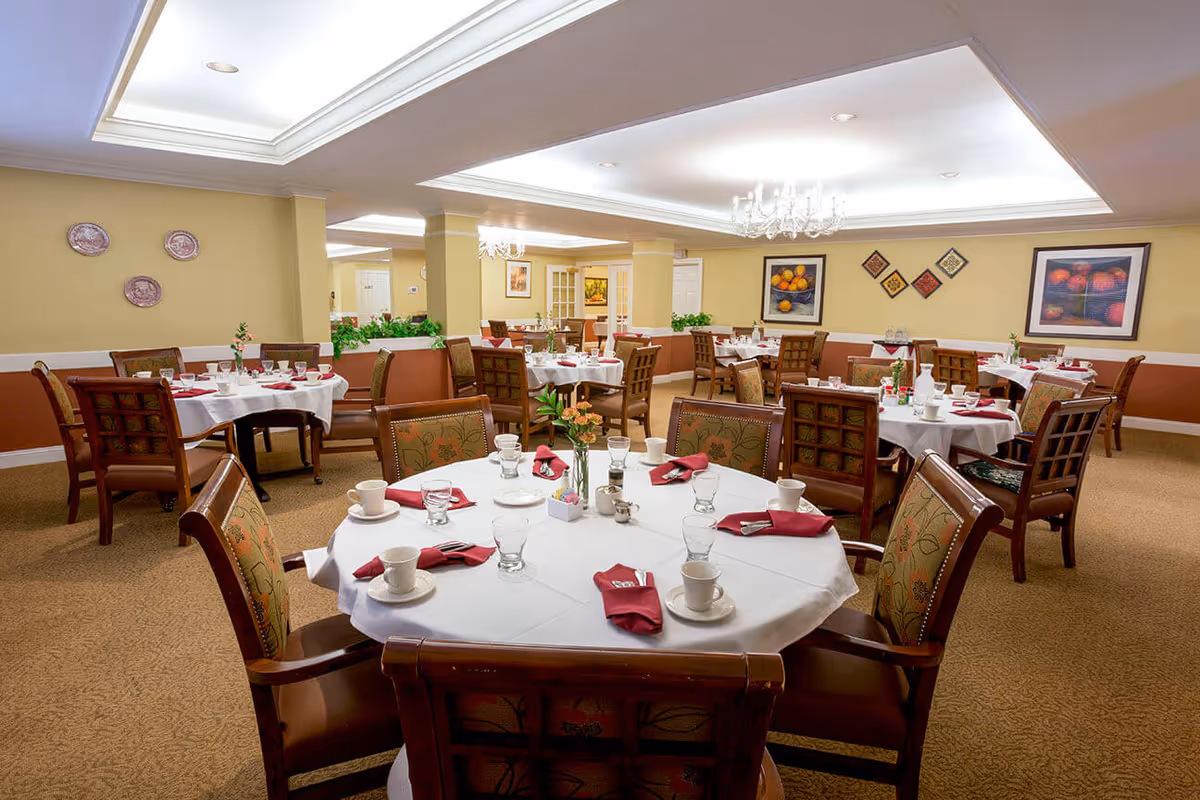 A well-lit dining room with multiple round tables covered with white tablecloths, each set with cups, glasses, plates, and red napkins. The chairs have wooden frames with floral upholstered backs and seats. The walls are painted beige with a white chair rail and decorated with framed artwork and decorative plates. The ceiling has recessed lighting and chandeliers.