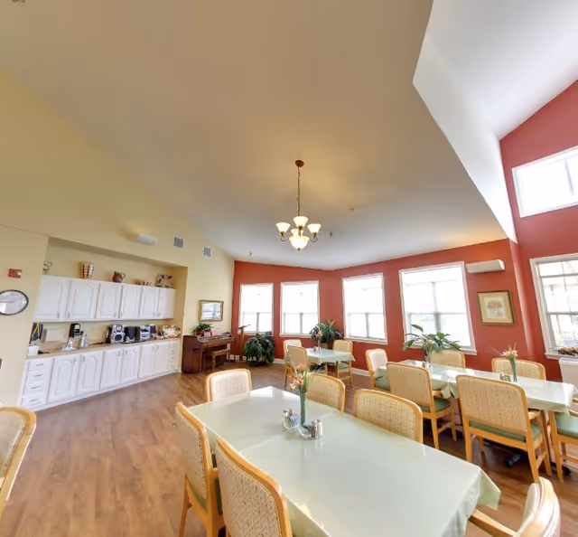 A bright dining room with several tables covered in light green tablecloths and surrounded by wooden chairs with woven backs. The room has large windows letting in natural light, red accent walls, and a wooden floor. There is a white cabinet with kitchen appliances along one wall and a chandelier hanging from the ceiling. Potted plants and framed pictures decorate the space.