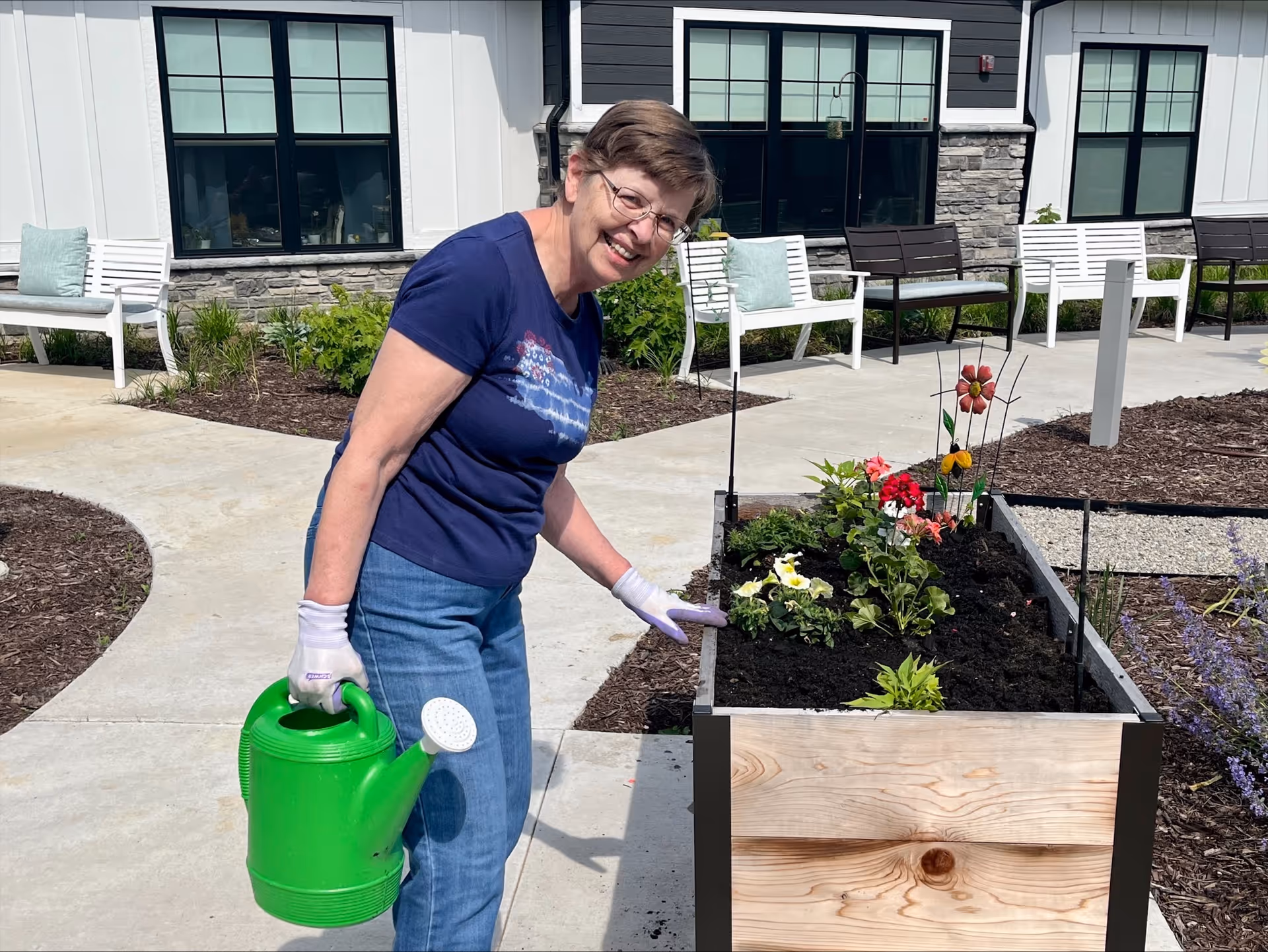 An elderly woman wearing glasses, a navy blue t-shirt, and jeans is watering plants in a raised garden bed outdoors. She is smiling and holding a bright green watering can. The background shows a paved walkway, garden beds, and benches in front of a building with large windows.