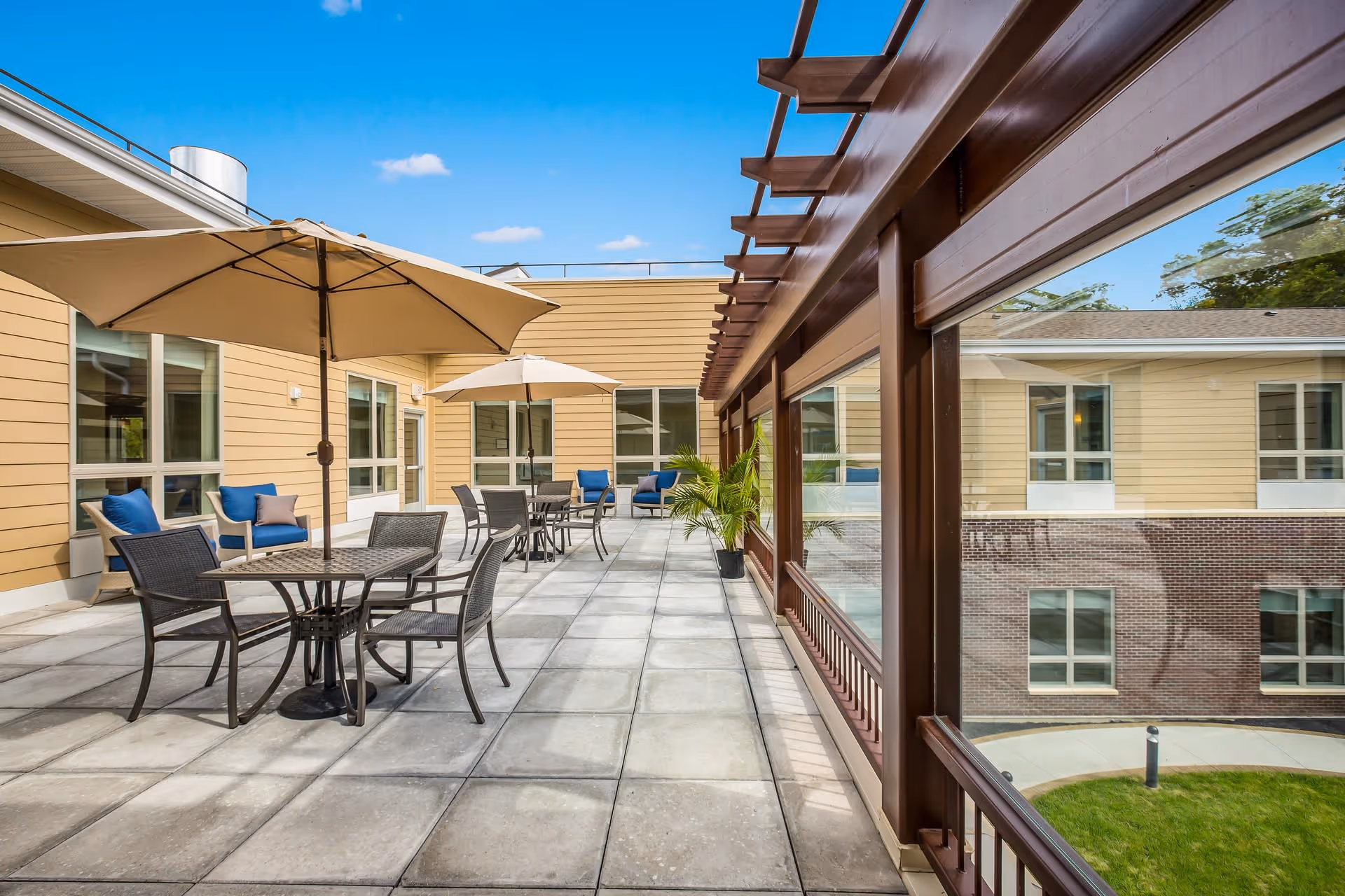 Sunny outdoor terrace with patio tables, umbrellas, chairs and lounge seating along a building courtyard.
