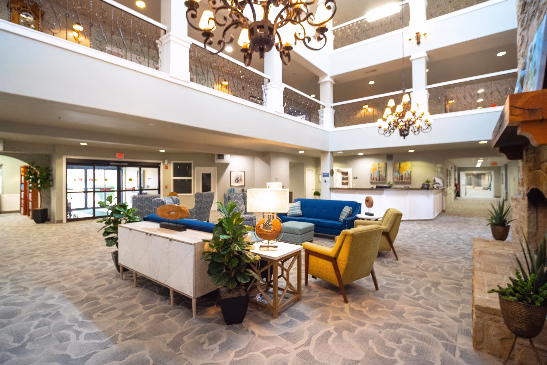 A spacious and well-lit common area in a retirement community featuring comfortable seating including blue sofas and yellow armchairs arranged around a central table with a lamp and decorative plants. The area has a high ceiling with ornate chandeliers, a stone fireplace on the right, and a balcony with decorative railings on the upper level. The floor is carpeted with a patterned design, and there are paintings and plants decorating the walls and corners.