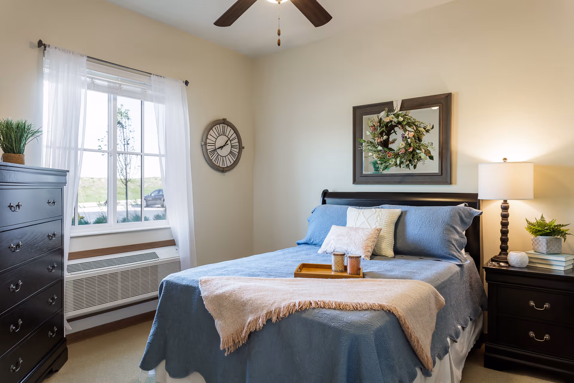 A cozy bedroom with a neatly made bed featuring blue bedding and decorative pillows. A beige throw blanket is draped across the foot of the bed with a wooden tray holding two candles. To the right of the bed is a dark wooden nightstand with a lamp, books, and a small potted plant. On the wall above the bed hangs a framed mirror with a floral wreath. To the left is a window with sheer white curtains, a wall clock, and a dark wooden dresser with a potted plant on top. The room has light-colored walls and a ceiling fan with dark blades.