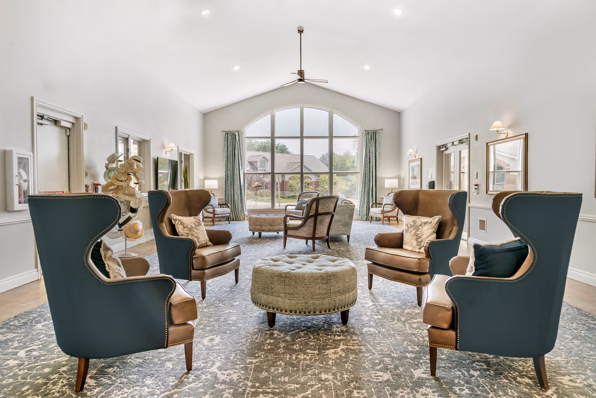 A bright and spacious living room with high vaulted ceilings and a large arched window letting in natural light. The room features four wingback chairs with cushions arranged around a round tufted ottoman on a patterned area rug. Additional seating and lamps are visible near the window, and the walls are painted white with simple wall sconces and framed mirrors.